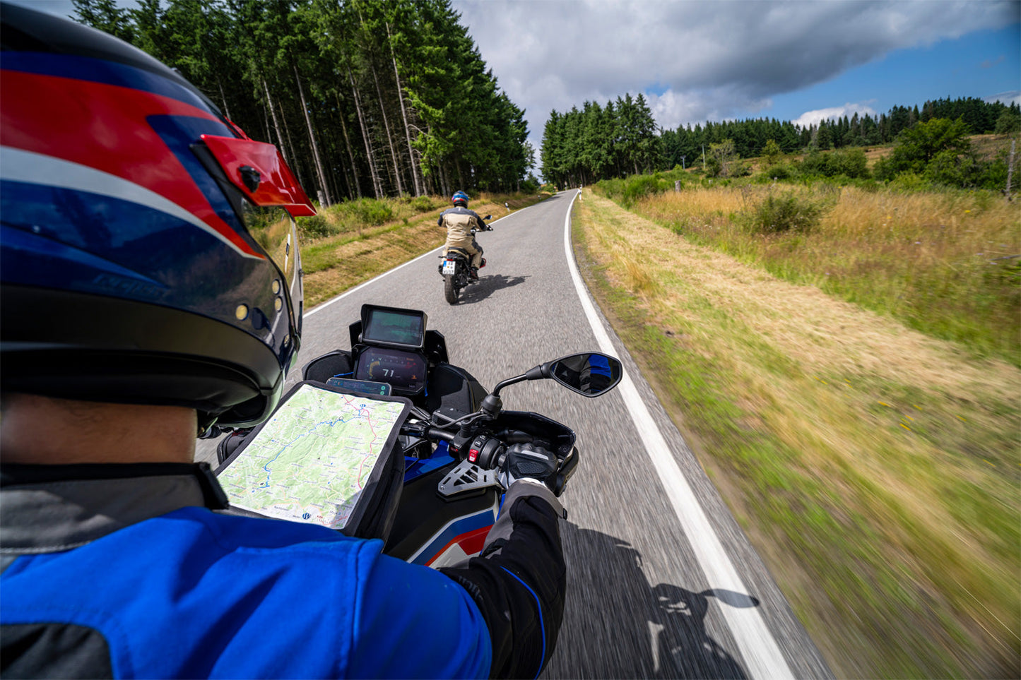 Two motorcyclists on a scenic road with trees and open fields.