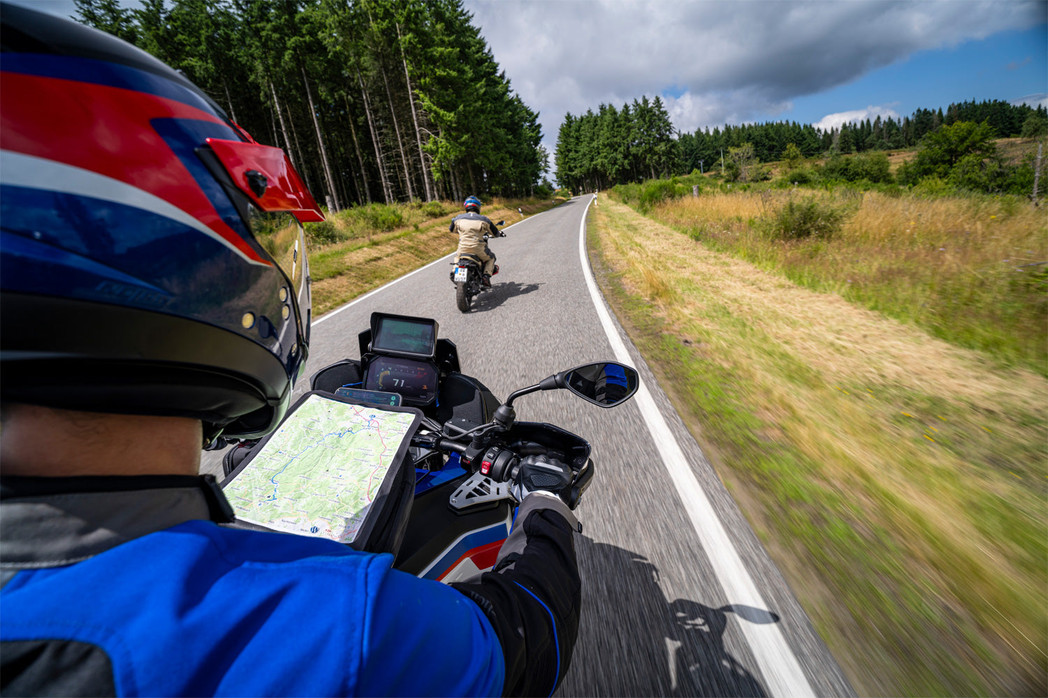Two motorcyclists on a scenic road with trees and open fields.